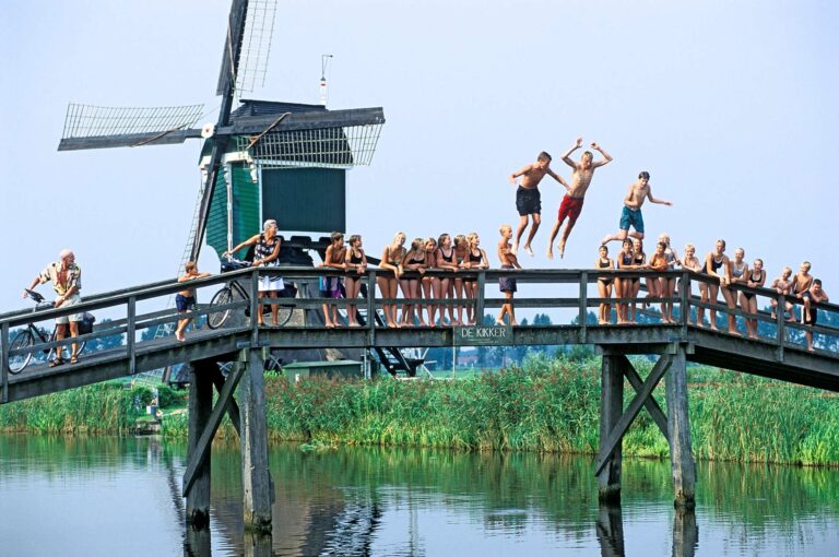 Kids jumping in water from a bridge in front of a windmill