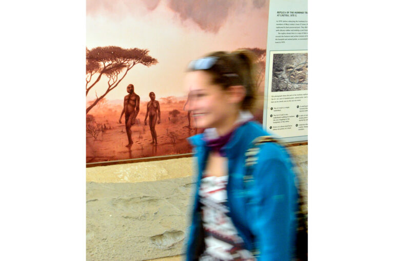 Een moderne vrouw loopt voor langs een afgietsel van de Laetoli voetafdrukken met tekening van onze voorouders, miljoenen jaren geleden. Olduvai Gorge Museum.