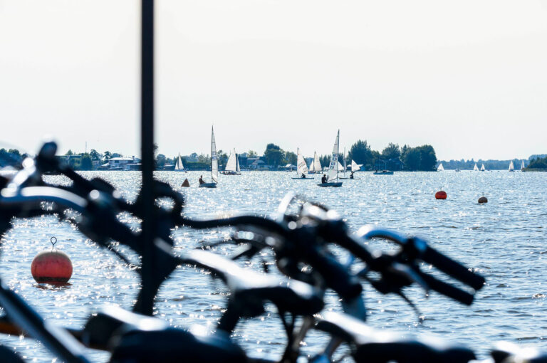 Fietsen op een boot op het water, met zeilbootjes in achtergrond.