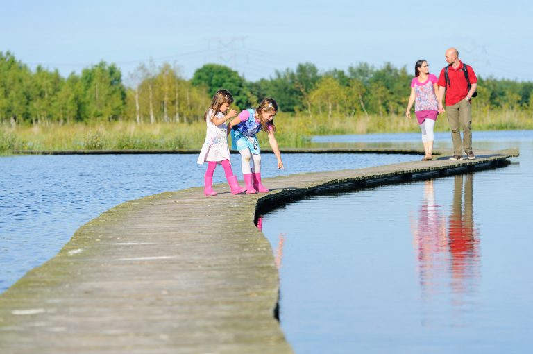 Gezin op vlonder over water in natuurgebied, de kinderen wijzen naar iets in het water en de ouders wandelen