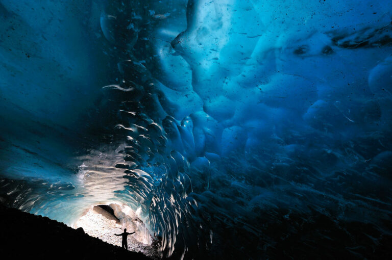 Een man staat met opgeheven armen in een ijsgrot onder een gletsjer