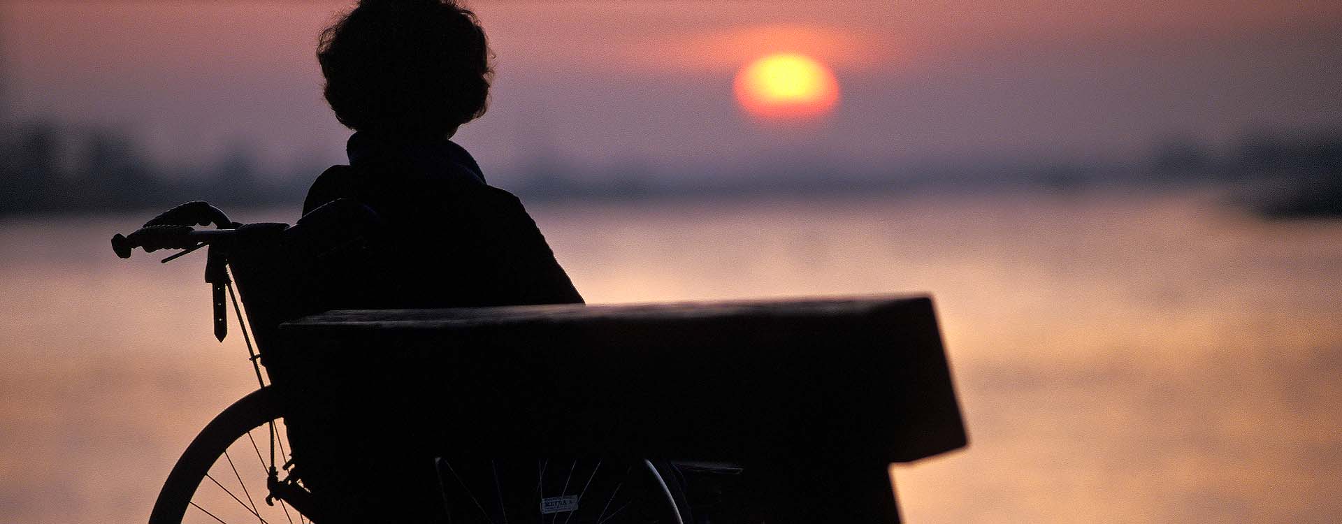 Mevrouw in rolstoel naast een bankje kijkt naar zonsondergang boven een rivier.