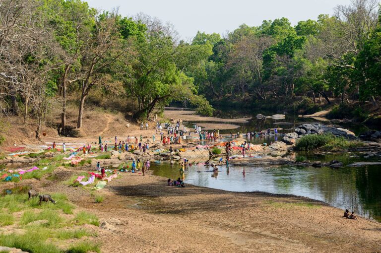 Public washing place with many people, along small water, a river, surrounded by forest.